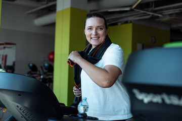 Happy overweight woman vigorously run on treadmill at fitness gym with towel hanging around neck,...