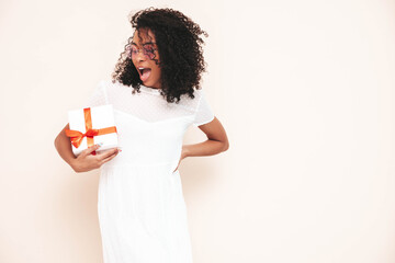 Beautiful black woman with curls hairstyle. Smiling model dressed in white summer dress. female posing near wall in studio. Tanned and cheerful. Holding gift box. Shocked, surprised