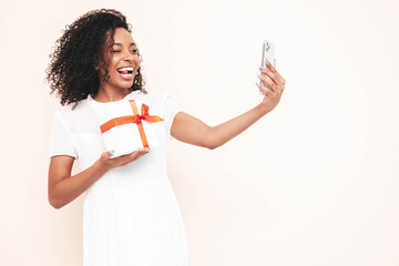 Beautiful black woman with curls hairstyle. Smiling model dressed in white summer dress. female posing near wall in studio. Tanned and cheerful. Holding gift box. Shocked, surprised. Take selfie