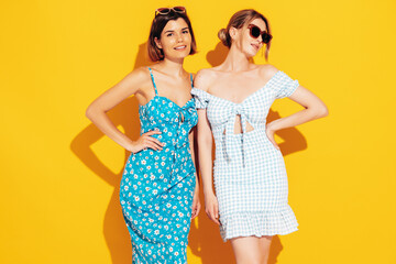 Two young beautiful smiling brunette hipster female in trendy summer dress clothes. Sexy carefree women posing near yellow wall. Positive models having fun. Cheerful and happy
