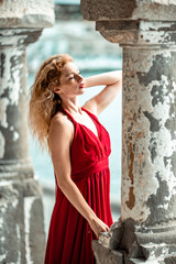 Obraz premium Outdoor portrait of a young beautiful natural redhead girl with freckles, long curly hair, in a red dress, posing against the background of the sea.