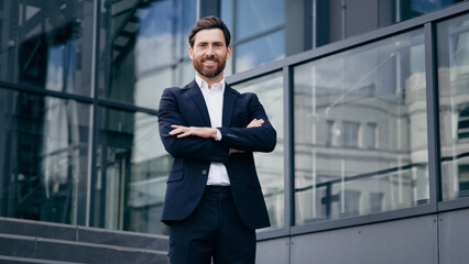 Successful confident caucasian bearded businessman in suit standing outdoors on background office building smiling proud male professional worker leader looking at camera posing with crossed arms - Powered by Adobe