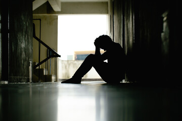 Silhouette of depressed man sitting head in hands on the walkway of residence building. Sad man,...