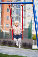 Little female child, wearing warm clothes, swaying on swing, having fun at playground. Front view of adorable girl on swing smiling, looking at camera, while swinging outdoors. Concept of joy.