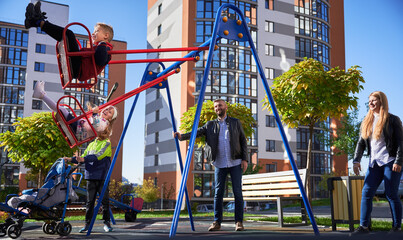 Family - father, mother and children having fun on playground. Parents swinging daughter and son, while other child standing with pram at modern courtyard of city residential high-rise buildings.