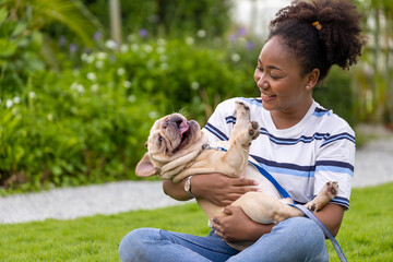 African American woman is playing with her french bulldog puppy while walking in the dog park at...