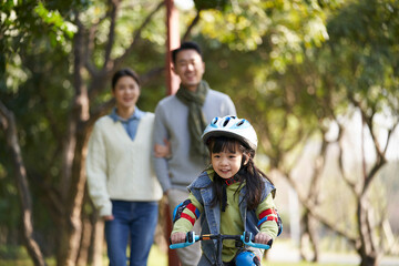 little asian girl riding bike in city park with parents in background