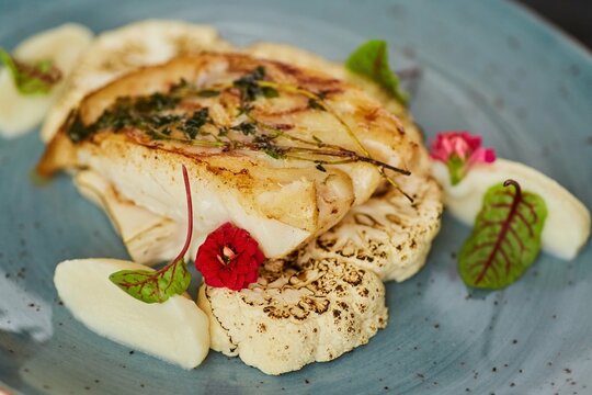 Sea or river baked white fish lies on a base of cauliflower vegetables with sauce. Decoration of flowers and herbs spices. A dish from the chef for serving