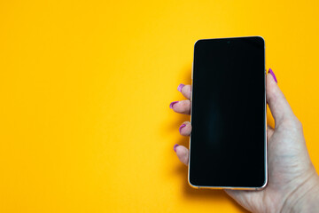woman hand with nails manicure holding smartphone with blank black screen on yellow background,...