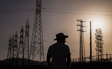Silhouette of adult man in cowboy hat against transmissions tower during sunset