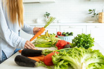 Woman take fresh carrot from table full of vegetables and greens in kitchen. Housewife make salad...