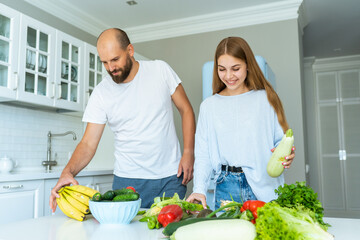 Joyful couple lay out fresh vegetables, fruits and greens on table in kitchen. Making salad for...