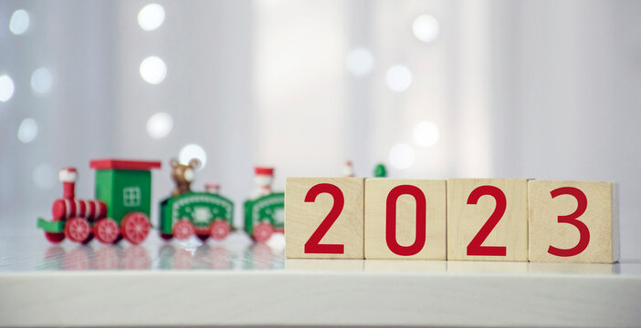 Coming soon New Year 2023 number on wooden cube block with colorful Christmas train and blurred bokeh lights on the background. Merry Christmas and Happy New Year greeting card for winter holidays.