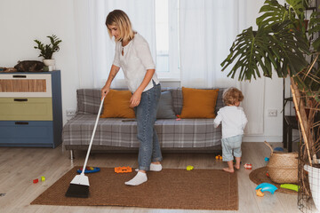 Young woman, housewife cleans up toys with broom together with her little son at home