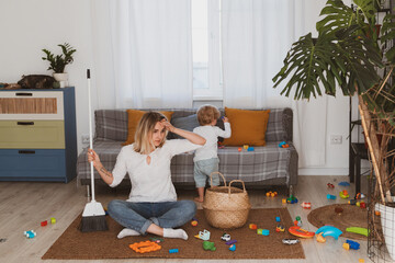 Young woman, housewife cleans up toys with broom together with her little son at home