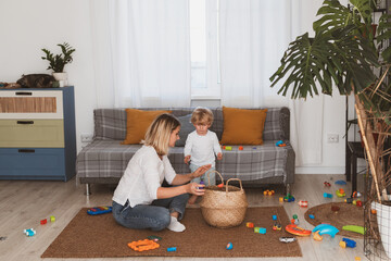 young woman, housewife puts toys in the basket with her little son at home