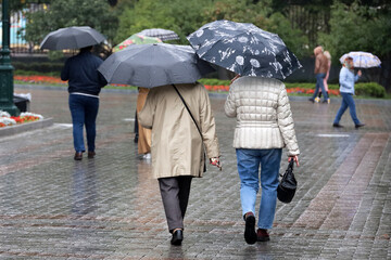 Rain in city, people with umbrellas walking on a street, two women in foreground. Rainy weather in autumn park