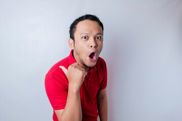 Shocked Asian man wearing a red t-shirt pointing at the copy space beside him, isolated by a white background