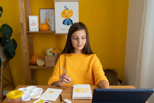 Smiling girl painting on sketch pad and watching online tutorial through tablet PC at home