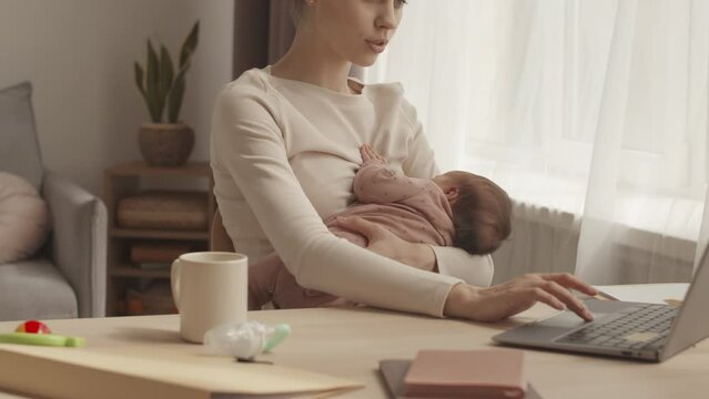 Waist up slowmo of young woman with infant baby daughter working on laptop from home, sitting at desk in cozy living room