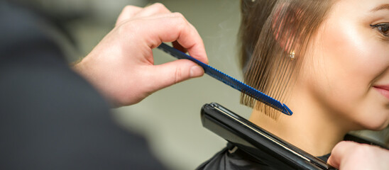 Hairstylist is straightening short hair of young brunette woman with a flat iron in a hairdresser...