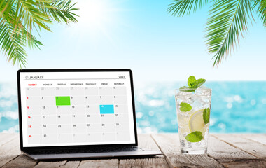 Laptop with calendar and cocktail on wooden table in front of sunny sea