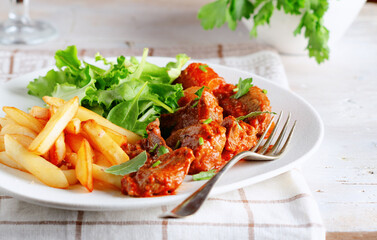 Close up of beef stew with herbs, chips and salad in dish on white wooden background.