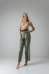 Attractive young woman with long straight blonde hair and natural makeup wearing crop top and casual trousers posing over grey background.
