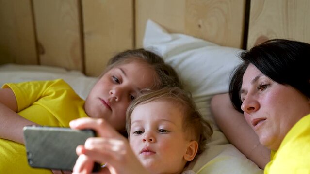 Happy family concept. Mom, daughter and son are play on bed. Child watching video on smartphone. Family leisure at home. Boy and girl with mom are watching movie on smartphone. Home education concept
