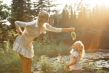 Side view of family standing near pond in park lit up by sunset in summer. Young woman holding...