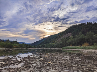 Fototapeta premium Beautiful cloudy sky at sunset over the river. The high bank of the river is overgrown with grass.