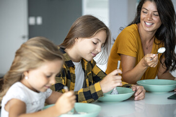 Teenage girl and little sister eating in kitchen with mom in background smiling and looking at them Focus on teenage girl