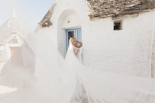 Portrait of young joyful woman bride with flying bridal veil, standing near old white stone house trullo in sunny Italy.