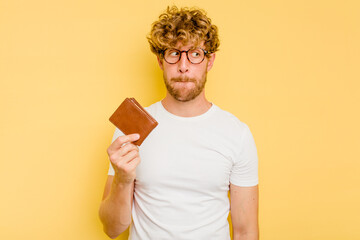 Young caucasian man holding a wallet isolated on yellow background confused, feels doubtful and...