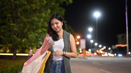Stylish woman typing text message on her mobile phone while walking through night city street with blurred lights background © Prathankarnpap