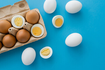 Top view of natural boiled and raw eggs near carton tray on blue background.