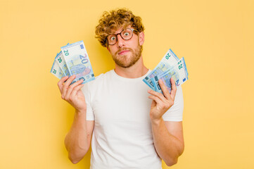 Young caucasian man holding a banknotes isolated on yellow background