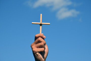 Small wooden cross holding in hand with cloudy and blue sky background, concept for love, hope, truth, faith, believe in Jesus, soft and selective focus.