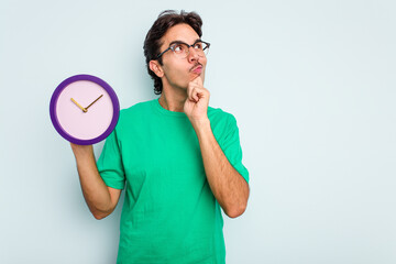 Young hispanic man holding a clock isolated on white background looking sideways with doubtful and skeptical expression.