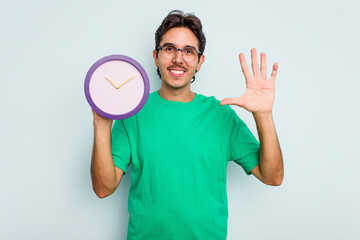Young hispanic man holding a clock isolated on white background smiling cheerful showing number...