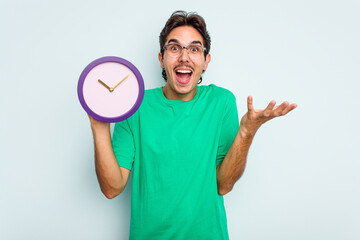 Young hispanic man holding a clock isolated on white background receiving a pleasant surprise, excited and raising hands.