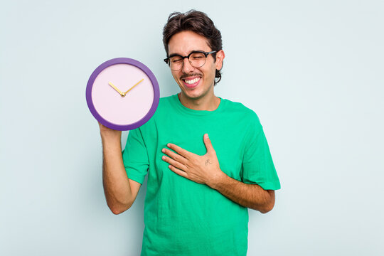 Young hispanic man holding a clock isolated on white background laughing and having fun.