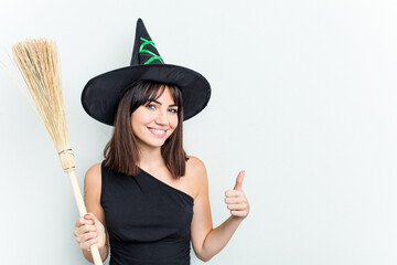 Young caucasian woman dressed as a witch holding a broom isolated on blue background smiling and raising thumb up