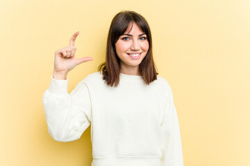 Young caucasian woman isolated on yellow background holding something little with forefingers, smiling and confident.