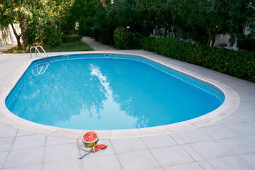 Half of a cut watermelon and a piece lie on a plate on the tiled floor by the pool. High quality photo