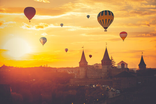 Colorful hot air balloons in orange sunset sky over ancient castle silhouette. Travel background. Natural summer scenery, evening sun dawn watching of old town cityscape. Kamianets-Podilsky, Ukraine