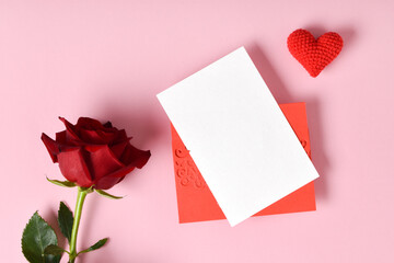 A red rose and a mock-up of a red envelope with a blank sheet on a pink background. Banner for Valentine's Day. Postcard for February 14. Love. Minimalism. A place to copy. Flat position, top view.