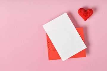 A mock-up of a red envelope with a blank sheet and a red knitted heart on a pink background.
