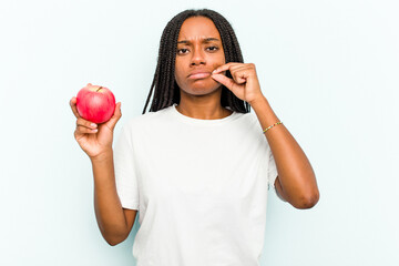 Young African American woman holding an apple isolated on blue background with fingers on lips keeping a secret.