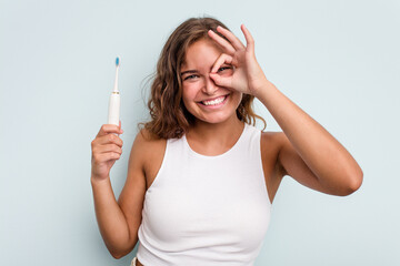 Young caucasian woman holding electric toothbrush isolated blue background excited keeping ok...